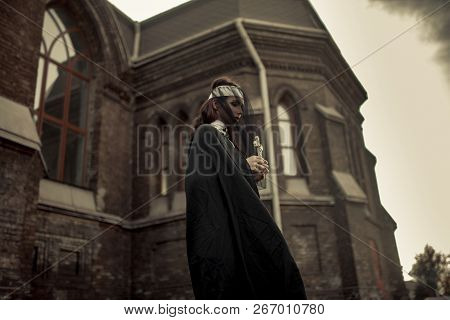 Beautiful Young Nun In Black Cassock, Cloak And Transparent Cape Is Standing And Praying With Cross 