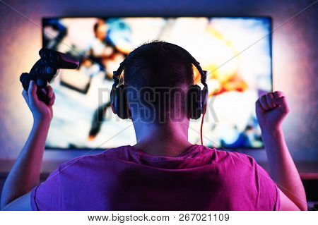 A Young Player Plays Video Games With Headphones, And A Gamepad At Home On A Large Tv.
