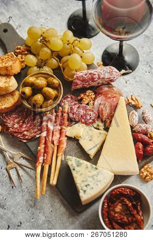 Antipasto platter cold meat and cheese board with grapes, wine, various kinds of cheese, grissini bread sticks on white rustic background. View from above