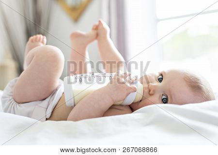 Pretty Baby Girl Drinks Water From Bottle Lying On Bed. Child Weared Diaper In Nursery Room.