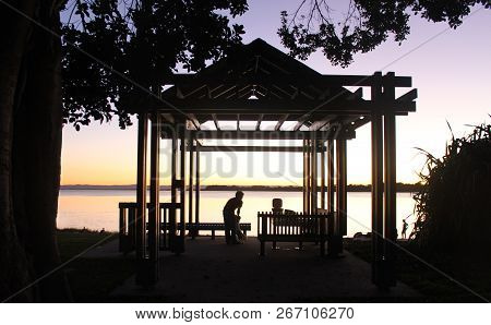 Pavilion By Water At Sunset - Silhouettes Of People And Dog With Overhanging Tree
