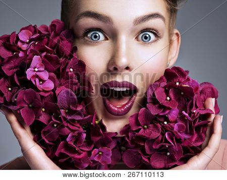 Closeup portrait of a surprised young woman. Female face with bright vivd makeup. Emotional girl with purple fowers.