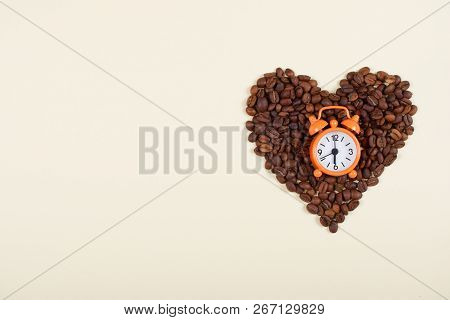 Roasted Brown Coffee Beans Laid In A Shape Of Heart With Orange Alarm Clock On A Beige Background Wi