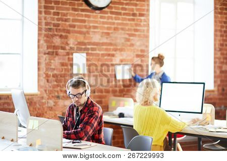 Serious young office worker in headphones looking at his notes while entering data in computer