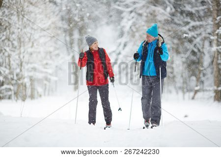 Active senior couple on skis moving down snowdrift in snowfall during training in winter forest