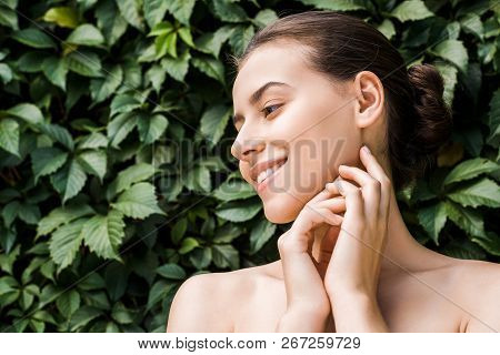 Smiling Young Woman With Hands Near Face On Green Leaves Background