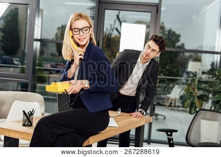 Businessman Looking At Young Businesswoman With Yellow Retro Telephone At Worklace In Office