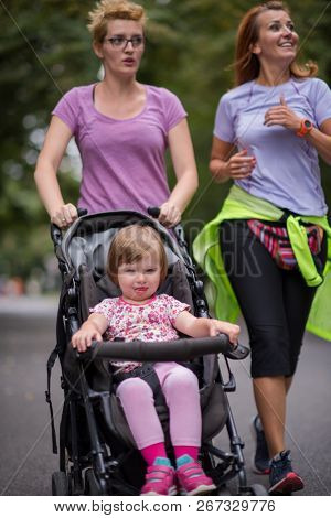 two young healthy women jogging together while pushing a baby stroller at city park
