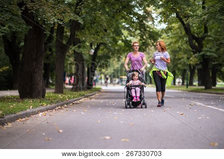 two young healthy women jogging together while pushing a baby stroller at city park
