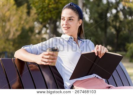 Woman Sitting On A Wooden Bench, Reading A Book And Drinking Coffee