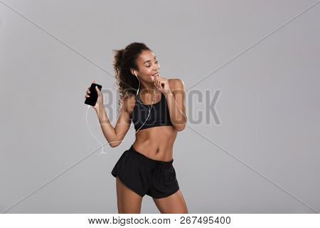 Portrait of an afro american happy sportswoman isolated over gray background, listening to music with earphones, holding mobile phone, dancing