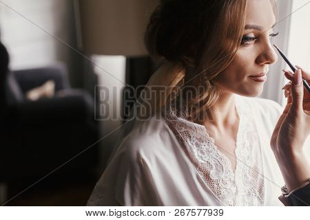 Stylish Bride Getting Her Make-up In The Morning At Window Light. Makeup Artist Doing Brows And Appl