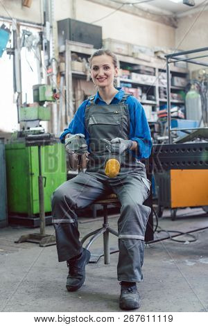 Female mechanic sitting in metal workshop looking and smiling into camera