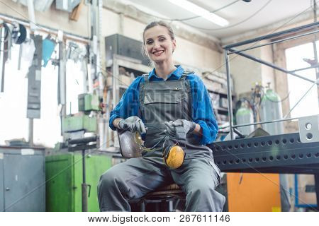 Female mechanic sitting in metal workshop looking and smiling into camera