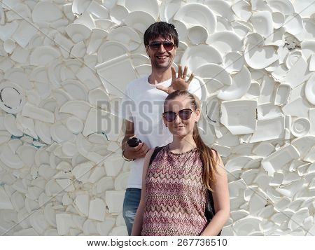 A Young Adult Guy Makes A Funny Sign Of The Crown Behind Caucasian Girl's Head. A Tall Dark-haired M