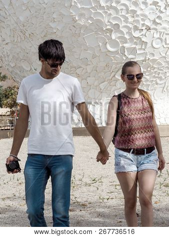 Young Adult Couple Of Lovers Go Holding Hands Against The Background Of A White Wall From Broken Dis