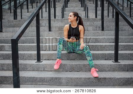 Well-built Young Woman Has Rest On Steps. She Sits There And Listen To Music Through Heaphones. Mode