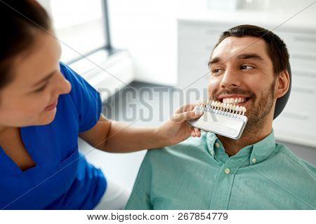 medicine, dentistry and healthcare concept - female dentist with tooth color samples choosing shade for male patient teeth at dental clinic