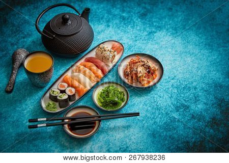 Various sushi rolls placed on ceramic plates. Traditional iron tea pot and green tea in cup. Kimchi and goma wakame salads. Soy souce and chopsticks on sides. Blue background with copy space.