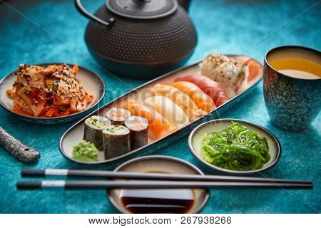 Various sushi rolls placed on ceramic plates. Traditional iron tea pot and green tea in cup. Kimchi and goma wakame salads. Soy souce and chopsticks on sides. Blue background.
