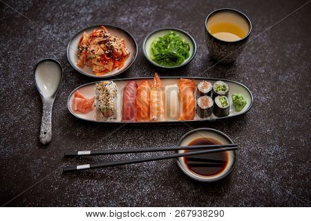 Asian food assortment. Various sushi rolls placed on ceramic plates. Kimchi and goma wakame salads. Soy souce and chopsticks on sides. Grungy dark background with copy space.