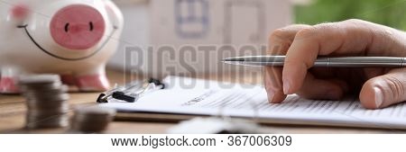 Man Fills In Lease Document At Home, On Piggy Bank. Composition And Value Property, Taking Into Acco