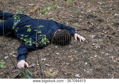 Murder In The Woods. The Body Of A Man In A Blue Shirt And Trousers Lies On The Ground Among The Tre