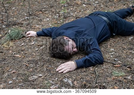 Murder In The Woods. The Body Of A Man In A Blue Shirt And Trousers Lies On The Ground Among The Tre
