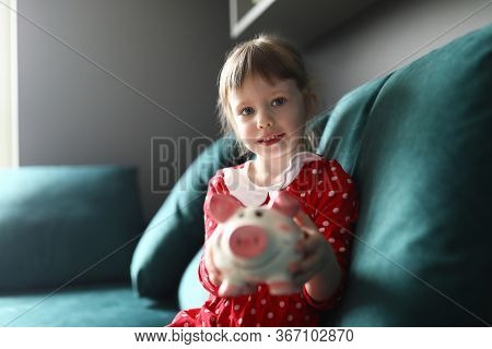Portrait Of Smiling Beautiful Child Holding Piggy Bank Sitting On Sofa Indoors. Lovely Blonde Kid Sa
