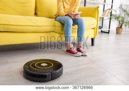 Partial View Of Child With Smartphone On Sofa Near Robotic Vacuum Cleaner On Floor In Living Room