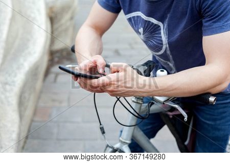 A Young Man Holding A Smartphone Sitting On A Bike Resting His Hands On The Handlebars. Biker / Cycl