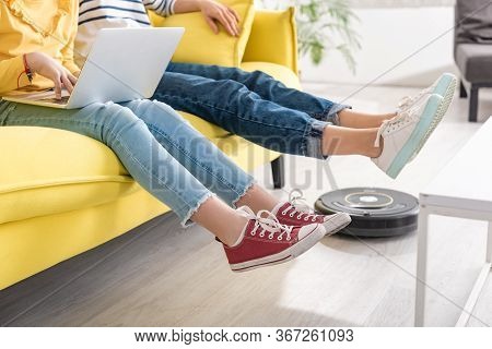 Cropped View Of Daughter With Laptop Near Mother With Raised Legs On Sofa And Robotic Vacuum Cleaner