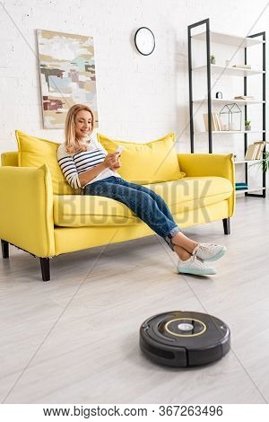 Beautiful Woman With Smartphone On Sofa Smiling And Looking At Robotic Vacuum Cleaner On Floor In Li