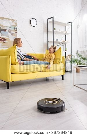 Mother And Daughter Looking At Each Other And Lying On Sofa Near Coffee Table And Robotic Vacuum Cle