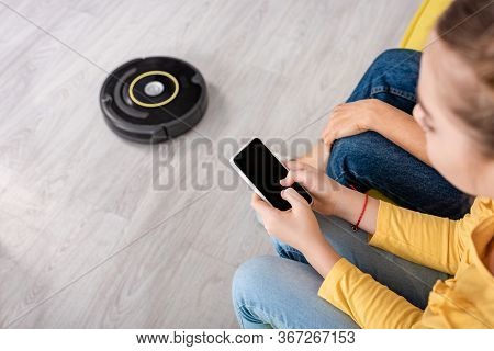 Cropped View Of Child With Smartphone And Mother Near Robotic Vacuum Cleaner On Floor In Living Room