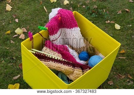 Knitting Needles, Knitting Threads And Knitted Clothes In A Yellow Box Standing On The Grass, Strewn