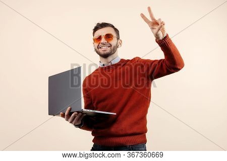 Positive Bearded Young Man In Casual Outfit With Notebook Or Computer Show Peace Gesture Over Beige 