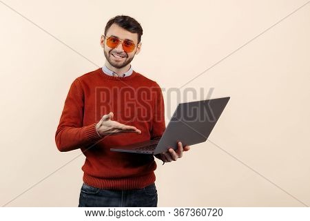 Joyful Bearded Young Man In Casual Outfit Show With Hand On Notebook Or Computer Over Beige Backgrou