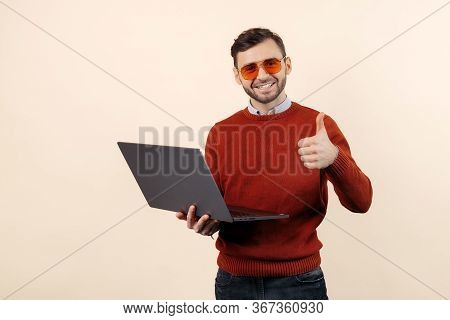 Handsome Bearded Young Man In Casual Outfit With Notebook Or Computer Show Thumbs Up Over Beige Back