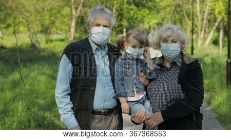 Old Grandparents With Granddaughter Together Walk In Spring Park In Medical Masks. Coronavirus Covid
