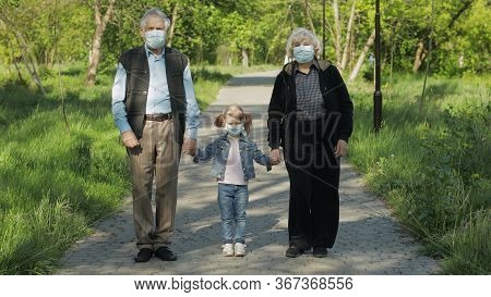 Old Grandparents With Granddaughter Together Walk In Spring Park In Medical Masks. Coronavirus Covid