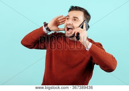 Shocked Bearded Young Man In Casual Outfit Talk On Smartphone Over Cyan Background.