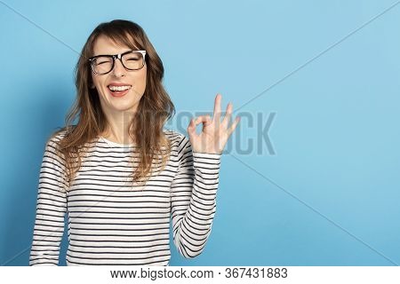 Portrait Of A Young Friendly Woman With A Smile In A Casual T-shirt And Glasses Makes An Okay Gestur