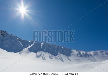 Sun In Blue Sky Shining On Mountain Scarp With Many Snowslide Deposits In Swiss Alps