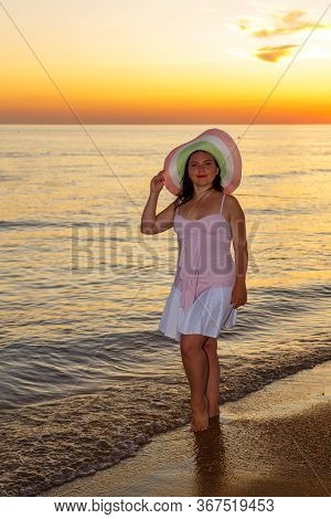 A Young Woman In A White Dress And Hat Stands On The Seashore At Sunset.