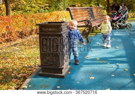 A Little Cute Baby In Blue Clothes Stands Near A Street Urn In A Park And Throws Garbage There And S