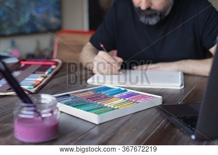 Man Artist Painting With Pencil And Pastel Crayon On Paper In Front Of Laptop. Learning Painting Onl