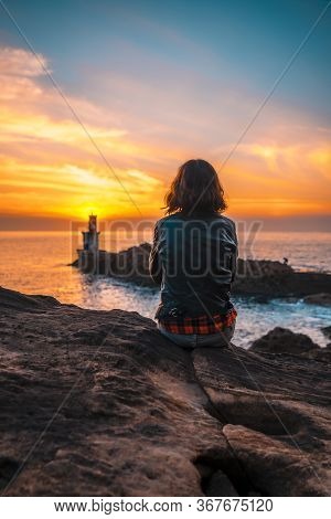 A Young Woman Sitting On Some Rocks With A Leather Jacket In The Orange Sunset Of The Pasajes San Ju