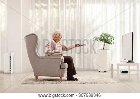 Elderly woman in an armchair changing channels on TV with remote control
