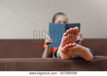 A Funny Little Caucasian Girl With Bare Feet With Book While Sitting On A Sofa At Home. Foot Closeup